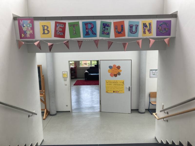 Ein Treppenabsatz mit einer bunten "Betreuung"-Girlande und einem Willkommensschild an einer Tür im Hintergrund.