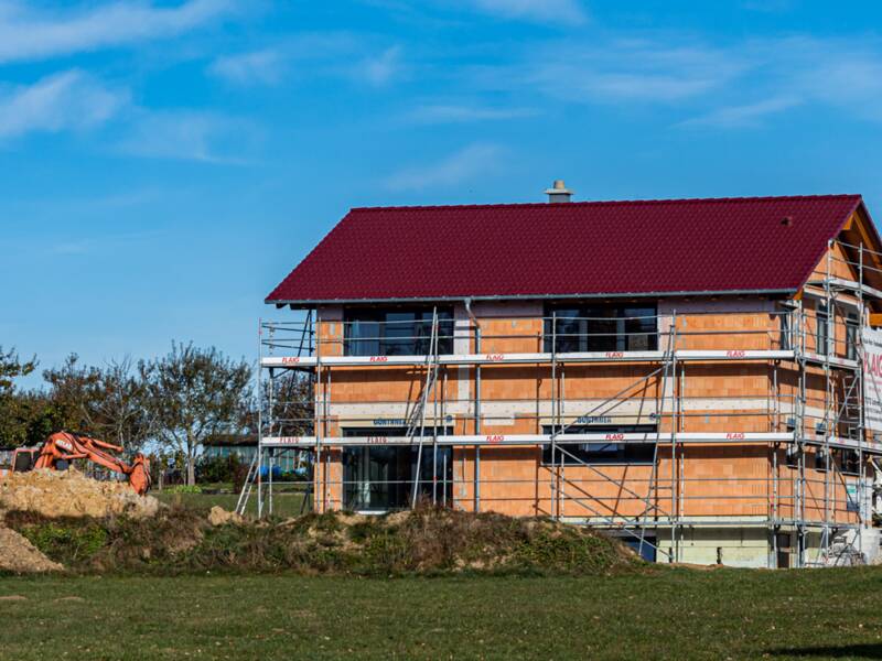 Neu gebautes Haus, das noch eingerüstet ist. Das Haus steht am Ende eines Wohngebiets auf einer grünen Wiese. Der Himmel ist blau.